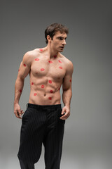 sexy man in black pants posing with red kiss prints on shirtless muscular torso and looking away on grey background.