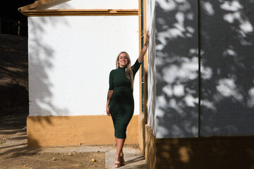 Obraz na płótnie Canvas Beautiful blonde woman with straight hair dressed in green dress. The woman is leaning on a happy white wall while the sun rays enter her back.