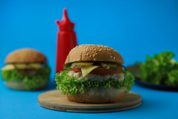 juicy delicious hamburger from fresh bun with sesame seeds, green salad, tomatoes, cucumbers, fragrant cheese and meat cutlets next to a bottle of ketchup in a red bottle
 on a bright blue background