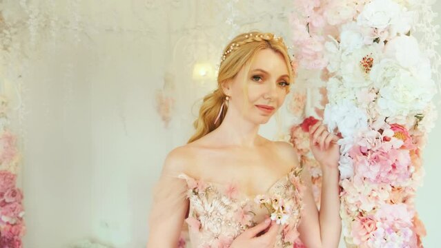 fantasy lady happy girl princess in white room spring scenery, flower decor. Woman queen blonde hair, vintage pink dress romantic hairstyle looking at camera fashion model posing, evening prom makeup