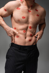 cropped view of man in black pants touching muscular torso with red lip prints isolated on grey.