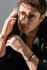 trendy man in black blazer and silver necklace with bracelet holding hand near face while looking...
