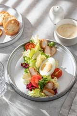 Caesar salad in modern linear feed. Healthy salad with chicken, egg, tomatoes, lettuce and parmesan chips with cutlery on gray table in harsh morning liht with window shadow. Light diet, menu