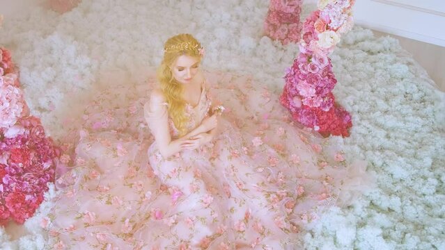 Fantasy happy woman fairy princess sitting on snow. magical fairy tale blonde hair queen. Girl smiling face. floral long pink evening dress creative design. Arch flowers decor. Lady goddess of spring