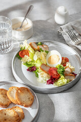 Caesar salad in modern linear feed. Healthy salad with chicken, egg, tomatoes, lettuce and parmesan chips with cutlery on gray table in harsh morning liht with window shadow. Light diet, menu
