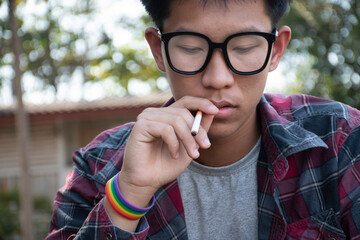 Young asian teen boy in plaid shirt wears rainbow wristband and holds cigarette in hand and smoking, blurred background, concept for bad behavior of young teens all over the world.