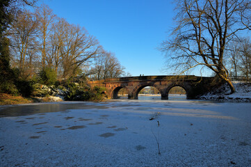 Obraz premium Eine winterliche Parklandschaft bei strahlend blauem Himmel und eine alte Sandsteinbrücke überspannt einen gefrorenen und schneebedeckten See