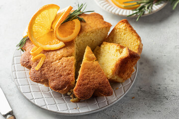 Sliced fluffy orange cake decorated with fresh orange slices on ceramic plate with knife on light gray textured background