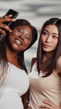 Vertical video: Two skincare models taking photos with smartphone, diverse young women advertising different skintones and body shapes. Beautiful luminous girls having fun with pictures for beauty ad.
