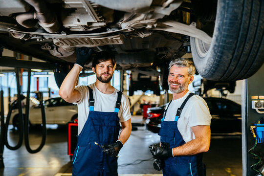 Two smiling mechanics, young and older men , stand under a car seen on a lift in a car service. Two car mechanic diagnosing vehicle at the auto service.