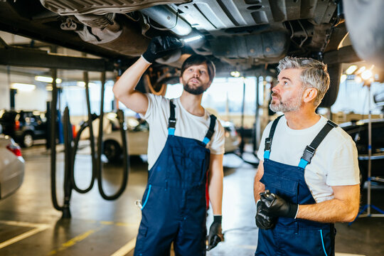 Two Auto Mechanics Men In Uniform At Car Suspension Repair Work. Vehicle Raised On Lift At Maintenance Station. Professionals Team Auto Mechanics Working On The Undercarriage Of A Car.