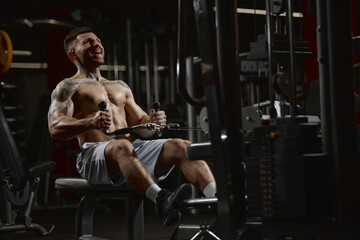 Fototapeta na wymiar Portrait of young muscular man training shirtless in gym indoors. Pulling rope with weigh. Relief body shape. Concept of sport, workout, strength