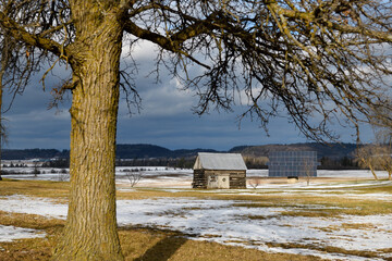 One room log cabin shack with solar panel in farmers field with snow clouds and naked tree in winter