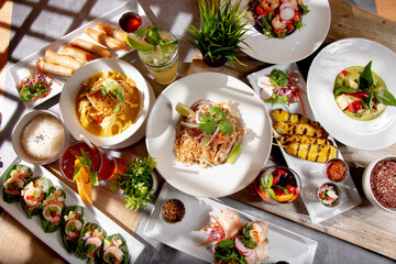 A top down view of a variety of Thai entrees on a dinner table, featuring pad Thai, Panang curry, chicken satay, green curry, and shrimp Miang Kham.