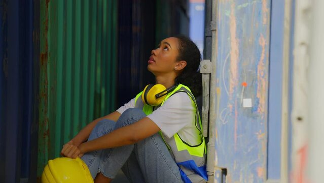 A african american female container yard worker wearing protective clothing and a hard hat sits tired next to the container after long hours of sweltering work in the berth.