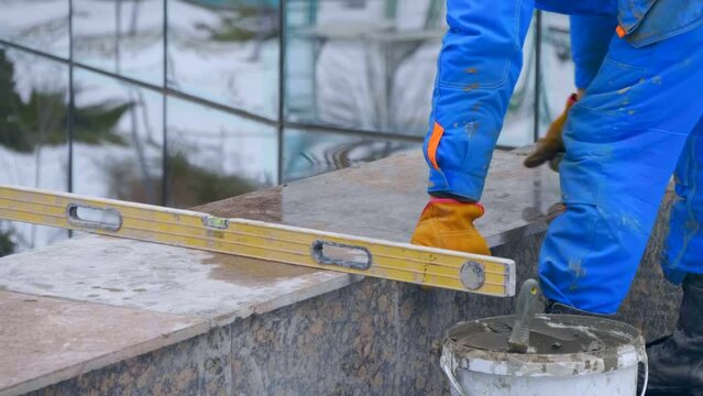 A worker lays on cement mortar and fine-tunes polished granite facing slab in a huge beautiful building complex under construction with glass fasade
