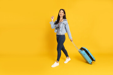 Happy Asian woman traveler walking and dragging a suitcase isolated on yellow background, Tourist girl having cheerful holiday trip concept, Full body composition