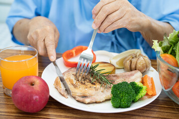 Asian senior or elderly old lady woman patient eating Salmon steak breakfast with vegetable healthy food while sitting and hungry on bed in hospital.