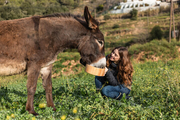 Woman smiling and holding bucket while feeding a donkey outdoors in nature.