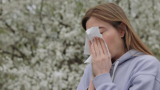 Unhealthy sick woman with allergy symptom blowing nose surrounded with seasonal flowering trees. Young caucasian lady having allergy symptoms outdoors