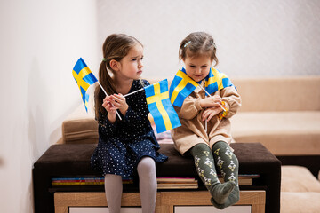 Two sisters are sitting on a couch at home with swedish flags on hands. Sweden children girls with...