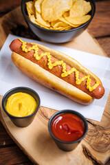 Classic fast food hot dog served with mustard on a rustic wooden board. Served with potato chips and sauces. 