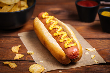 Classic fast food hot dog served with mustard on a rustic wooden board. Served with potato chips...
