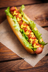 Hot dog with mustard, ketchup and green salad in bun, served with potato chips on a rustic wooden board. Fast food and delicious.