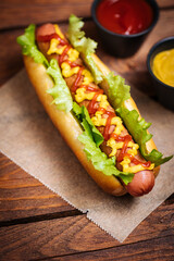 Hot dog with mustard, ketchup and green salad in bun, served with potato chips on a rustic wooden board. Fast food and delicious.