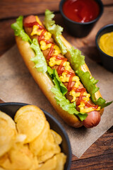 Hot dog with mustard, ketchup and green salad in bun, served with potato chips on a rustic wooden board. Fast food and delicious.