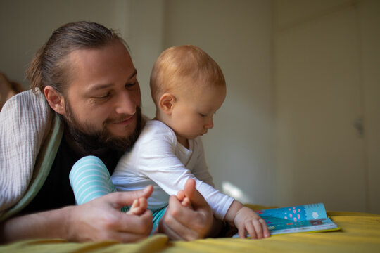 Dad with a beard and long hair reads a book to a kid.