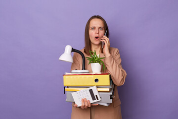 Photo of shocked astonished Caucasian woman wearing official style jacket holding folders isolated over purple background, hearing shocked news while talking cell phone.