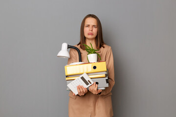Indoor shot of stressed sad woman wearing stylish beige jacket holding office stuff posing isolated...