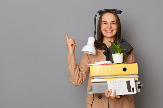 Portrait of funny young pretty woman wearing beige jacket holding documents and her office stuff posing with phone on her head isolated over gray background, showing advertisement area.