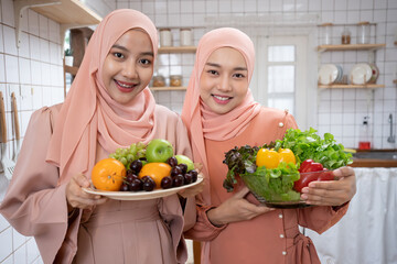 Happy young Asian woman friends couple standing in front of kitchen counter, holding salad in glass bowl and looking at each other with sweet smile while cooking together in kitchen at home.