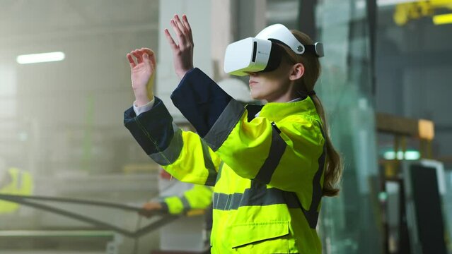 Portrait of Caucasian female manager in headset and yellow uniform standing in manufactory. Virtual reality experience. Engineering production concept. Indoor. Woman in VR goggles moving hands in air.