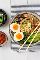 Bowl of ramen soup with chicken breast, vegetables, mushrooms and egg on a striped napkin on a white wooden table and chopsticks. Top view, healthy eating concept