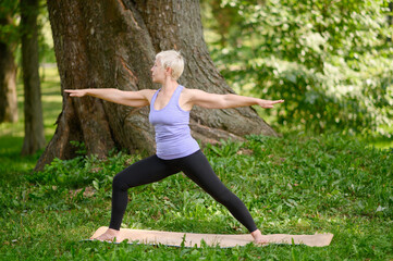 Middle aged caucasian woman in sportswear doing yoga outdoors in warrior pose, Virabhadrasana II. The concept of yoga, stretching, healthy lifestyle. Sports activities in nature. Copy space. 