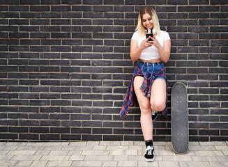 Happy young cheerful millennial skateboarder student girl standing in front of a black brick wall and chatting on her smartphone - People lifestyle and technology concept with teenager