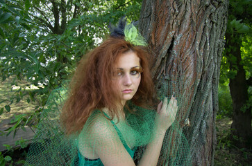 A girl in the form of a forest nymph near a tree against the background of tree bark and green foliage.