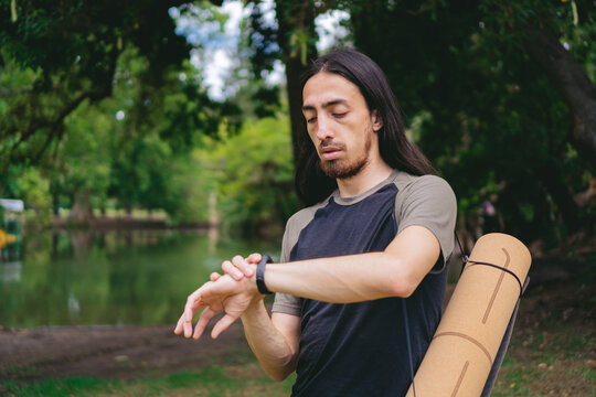 Young latin man with long hair in a forest with lake interacting with his smart watch.