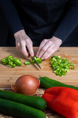 Hands of working woman cutting green pepper on a wooden kitchen board. Decofused gray bowls with eggs, red and green peppers, onion and courgette. Food preparation concept.