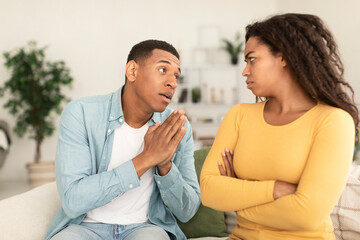 Sad young african american man asking for forgiveness from angry offended female in living room interior