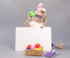 Kid looking at easter egg and hiding behind white board over gray background. Child wearing bunny ears. Space for design