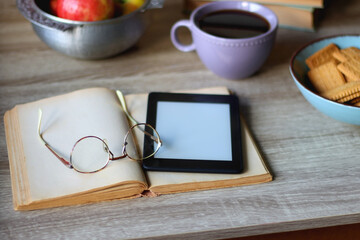 Stack of books, e-reader, reading glasses, bowl of biscuits, apples and cup of tea on the table. Bookshelf in the background. Selective focus.