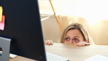 Woman hides behind office desk and looks at computer monitor.