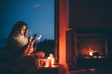 Cozy home. Young woman using phone near the fireplace.