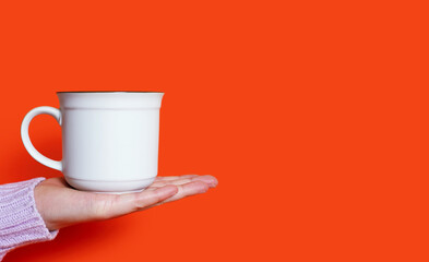A woman's hand holds a white cup with a hot drink on an orange background. The hand of a woman dressed in a pink sweater holds a ceramic cup against a bright orange background. Free space for text