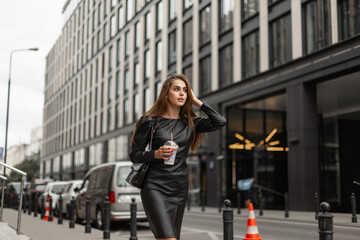 Fashionable beautiful business woman in fashion black clothes with a stylish dress and a leather bag with a cup of coffee walks in the city near a modern office building. Urban pretty girl
