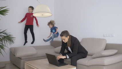 Young business woman working on laptop, children play and jump on sofa, stress,